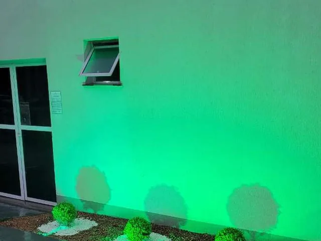 Fachada iluminada em verde à noite com canteiro e arbustos recém-plantados