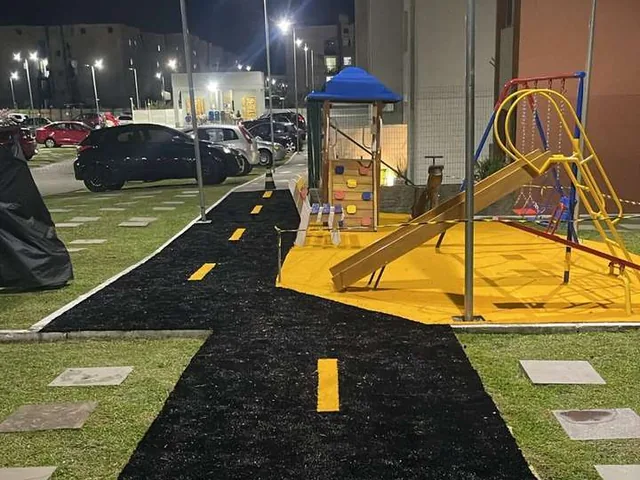 Praça do condomínio à noite com piso asfáltico novo, faixa de pedestres pintada e playground